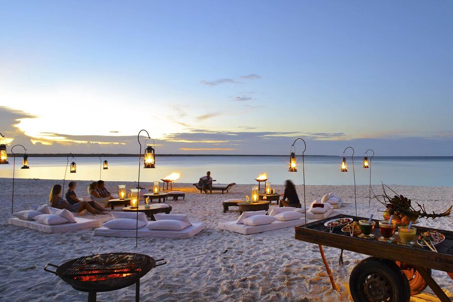 Guest enjoying drinks on the beach with ocean and meat grilling at sunset at andBeyond Mnemba Island.